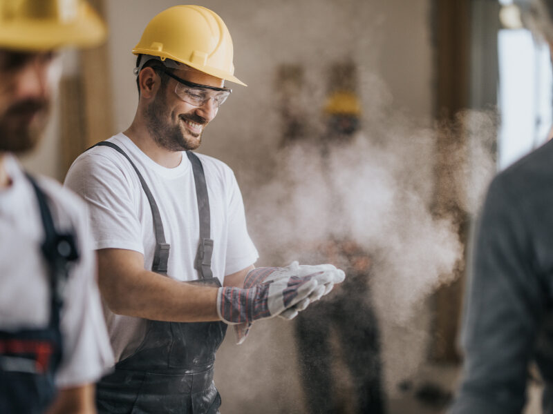 Young happy manual worker cleaning sawdust from his protective gloves at construction site.