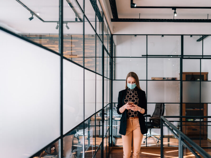 Young professional woman with mask on, walking through modern office, looking at her phone.